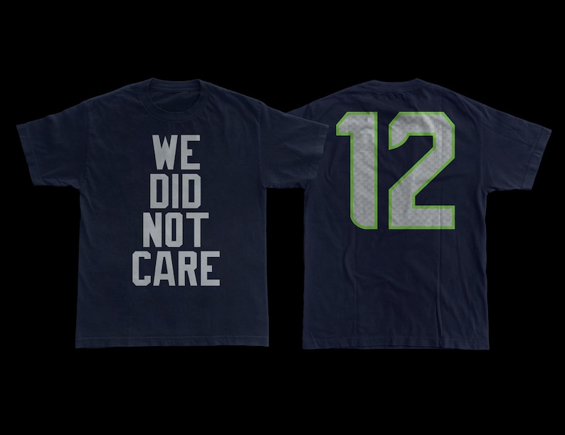 May include: Navy blue t-shirt with the text "WE DID NOT CARE" on the front. The back of the shirt displays the number "12" in a silver and green design. The shirt is made of a soft material.
