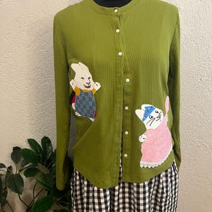 May include: A long-sleeved, ribbed olive green cardigan featuring a button-down front. The garment is decorated with two fabric appliqués: a white rabbit wearing blue overalls on the left and a white cat wearing a pink dress on the right.