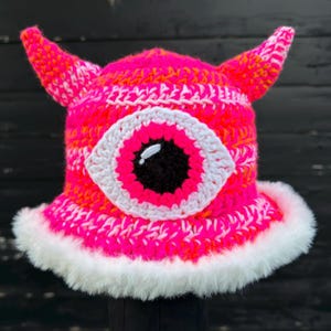 May include: A neon pink and orange crochet bucket hat featuring two pointed horns on top and a large central eye motif. The eye consists of a black pupil, a neon pink iris, and a white border. The brim is trimmed with thick white faux fur.