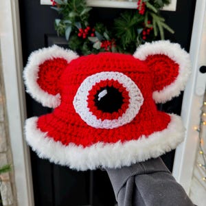 May include: A handmade red crochet hat with a large white-rimmed eye design and fluffy white trim. The hat has ear-like protrusions and is likely a novelty item or costume accessory.