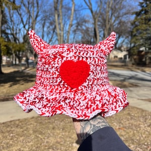 May include: A red and white crochet bucket hat with devil horns and a red heart. The hat is handmade with a ruffled brim. The hat is held up in front of a blurred outdoor background.