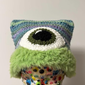 May include: A handmade crochet hat with a large, central eye design. The eye features a black pupil, olive green iris, and white sclera. The hat has a blue, green, and purple striped pattern and a fluffy green trim.