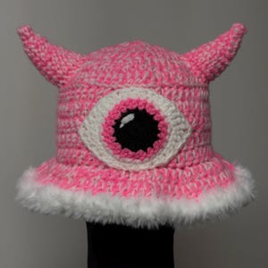 May include: A handmade pink crochet hat with white accents and two small horns. The hat features a large eye design with a black pupil, pink iris, and white sclera. The brim is trimmed with white faux fur.
