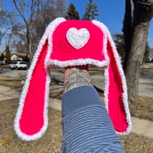 May include: A pink crocheted hat with white trim and a white heart on the front. The hat has long, floppy ears that are also crocheted.