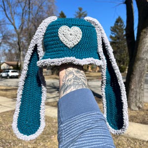 May include: Teal blue crochet hat with long, floppy ears and a white heart on the top. The hat is trimmed with a white fuzzy border.