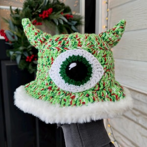 May include: A handmade crochet hat in green, red, and white yarn, featuring a large, white-rimmed eye with a green iris and black pupil. The hat has two pointed horns and a white faux fur trim around the brim. A whimsical, festive accessory.