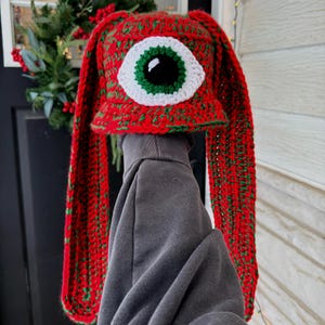 May include: A red and green crocheted hat with long ear flaps, featuring a large white-rimmed eye with a green iris and black pupil. The hat is handmade and has a festive, whimsical design.