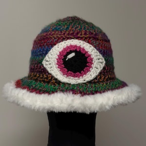 May include: A handmade crochet bucket hat with a large eye design. The hat features a multicolored yarn with a white, pink, and black eye. The brim is trimmed with white faux fur. This unique hat is a fun accessory.