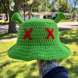 May include: Green crocheted bucket hat with red embroidered X's on the front and two green crocheted ears on top.