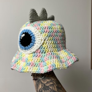 May include: A handmade crochet bucket hat in pastel colors, featuring a large eye design with a blue iris and black pupil. The hat has a gray, three-pointed crown and a wide brim. The hat is a unique, whimsical accessory.