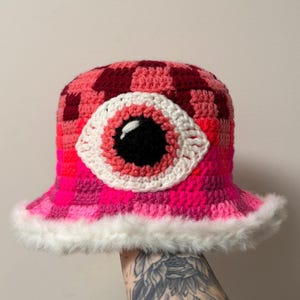 May include: A handmade crochet bucket hat in a vibrant pink and burgundy checkerboard pattern. The hat features a large, white-rimmed eye with a black pupil. The brim is trimmed with fluffy white faux fur, adding a playful touch to this unique accessory.