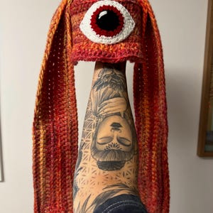 May include: A crochet hat in red, orange, and yellow, with long ear flaps. The hat features a large, embroidered eye with a white border, a red iris, and a black pupil. The hat is handmade. The hat is held up.