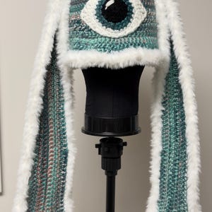May include: A handmade crochet hat with a large eye design and long, fluffy white ear flaps. The hat features a mix of teal, pink, and gray yarn, with a white and black eye. The ear flaps are trimmed with white faux fur.