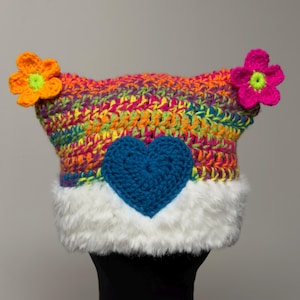 May include: A colorful crochet hat with a blue heart and two flower accents. The hat features a rainbow of colors, including orange, yellow, pink, and green. The brim is trimmed with white faux fur.