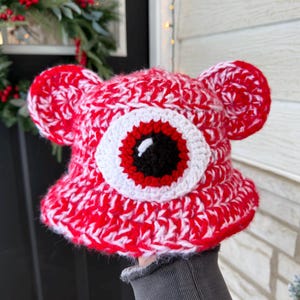 May include: A red and white crocheted hat with a large eye design. The hat features a black pupil, a red iris, and a white sclera. The hat has two ear-like protrusions on the sides.