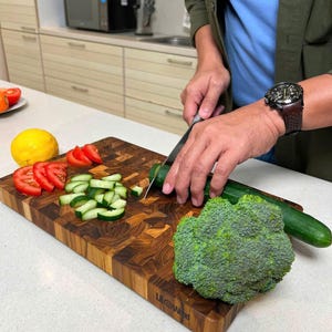 May include: A rectangular end-grain wooden cutting board with a dark brown checkered pattern. The board features a visible brand logo on the front edge and holds sliced red tomatoes, diced green cucumbers, a whole lemon, and a head of broccoli.