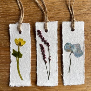 Könnte beinhalten: Drei handgefertigte Papier-Lesezeichen mit getrockneten Blumen und Bindfaden. Ein Lesezeichen zeigt eine gelbe Blume, ein anderes lila Zweige und das dritte eine blaue Blume. Jedes Lesezeichen ist rechteckig mit einer natürlichen Bindfadenschlaufe.