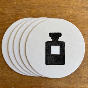 Könnte beinhalten: Sechs runde, cremefarbene Untersetzer. Jeder Untersetzer zeigt eine schwarze Silhouette einer Parfümflasche mit einem weißen rechteckigen Etikett. Die Untersetzer sind auf einer Holzoberfläche angeordnet.