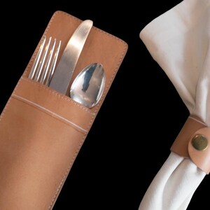 May include: A tan leather cutlery holder containing a silver fork, knife, and spoon, alongside a white fabric napkin secured with a matching tan leather napkin ring featuring a single brass-colored metal snap button closure.