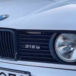 May include: Close-up of a white BMW 318is car with a black grille and a round headlight. The car has a silver emblem on the hood.