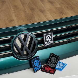 Peut inclure: Une calandre de voiture Volkswagen vert foncé avec un logo circulaire noir mat et un emblème rectangulaire noir et blanc Volkswagen Motorsport. Quatre petits emblèmes rectangulaires supplémentaires en bleu, noir, rouge et bleu clair sont placés en dessous.