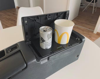 May include: A black car console with a built-in cup holder. A silver can with the text "The Coca-Cola Edition" and a white paper cup with the McDonald's logo are in the cup holder.