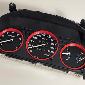 May include: A black car dashboard with three gauges. The gauges have red bezels and white needles. The speedometer reads up to 220 km/h. The tachometer reads up to 8,000 rpm. The fuel gauge is full. The temperature gauge is at the normal operating temperature.