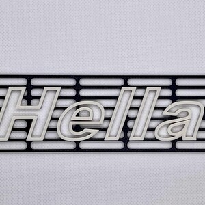 Puede incluir: Una rejilla rectangular negra con la palabra "Hella" en letras blancas estilizadas. La rejilla tiene ranuras horizontales y orificios de montaje. El diseño es un artículo impreso en 3D.