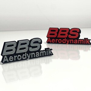 Könnte beinhalten: Zwei schwarze Plastikschilder mit dem Text "BBS Aerodynamik" in erhabenen Buchstaben. Ein Schild hat silberne Buchstaben und das andere rote Buchstaben.