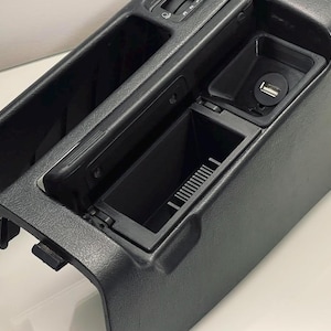 Könnte beinhalten: Schwarze Kunststoff-Mittelkonsole für Autos mit einem Ablagefach, einem kleinen offenen Fach und einem USB-Anschluss.