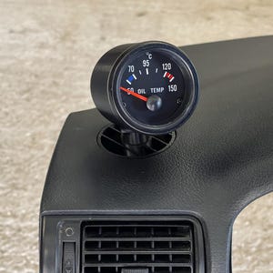 Puede incluir: Un manómetro de temperatura de aceite negro con una aguja roja que apunta a 70 grados Celsius. El manómetro está montado en un tablero negro con una ventilación debajo.