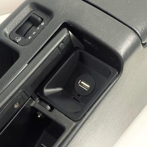 Puede incluir: Un panel interior de coche de plástico negro con un puerto USB, un interruptor de luz y un compartimento de almacenamiento.
