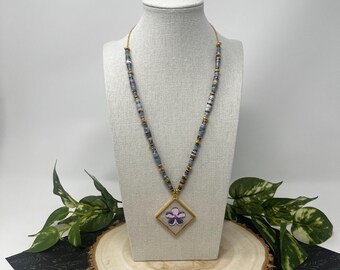 May include: A necklace with a gold chain and a pendant featuring a purple flower inside a gold diamond-shaped frame. The necklace has a beaded design with blue, gray, and gold accents. The necklace is displayed on a mannequin.