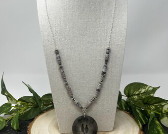 May include: A silver chain necklace with a large, round, dark gray pendant featuring a silhouette of two birds. The necklace has gray and silver beads and is displayed on a mannequin.