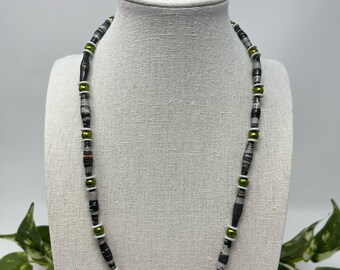 May include: A 20-inch necklace displayed on a mannequin. The necklace features a pattern of black, white, and gray beads, interspersed with olive green and white beads. The necklace is a handmade accessory.