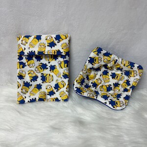 Può includere: Una pochette in tessuto bianco con un bordo blu e un motivo di Minion gialli. La pochette è aperta e un quadrato di tessuto bianco abbinato con un bordo blu e un motivo di Minion è all'interno.