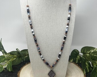 May include: A beaded necklace with a silver chain and a diamond-shaped pendant. The necklace features a mix of black, white, and blue beads, interspersed with copper-colored spacers. The pendant has a swirling design in shades of gray and purple.