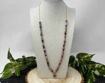 May include: A colorful necklace with a gold chain and a pendant. The necklace features a variety of small, rectangular beads in shades of red, blue, yellow, and purple. The pendant is a small, round charm with a pink center.