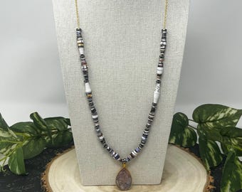May include: A handcrafted necklace featuring a gold chain and a pendant with a teardrop-shaped stone. The necklace has a beaded design with white, black, and gray beads. The pendant is a light purple color with gold accents.