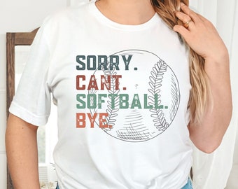 May include: White t-shirt with the text "SORRY. CAN'T. SOFTBALL. BYE." in a mix of colors. A sketched softball graphic is behind the text. The shirt is a casual style, suitable for sports enthusiasts.