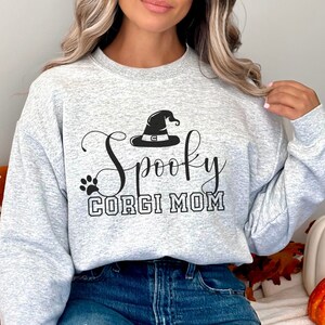May include: A gray sweatshirt with the text "Spooky Corgi Mom" in black lettering. A black witch's hat with a buckle is above the text. A black paw print is to the left of the text.