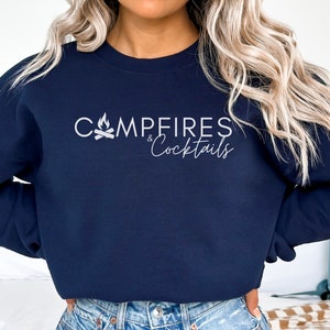 May include: A navy blue sweatshirt with the text "Campfires & Cocktails" in white lettering. A campfire icon is above the text.