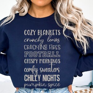 May include: A navy blue sweatshirt with a white graphic print that reads "Cozy Blankets, Crunchy Leaves, Crackling Fires, Football, Crispy Mornings, Comfy Sweaters, Chilly Nights, Pumpkin Spice."