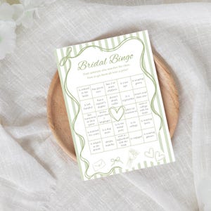 Può includere: Una carta da gioco rettangolare Bridal Bingo con uno sfondo a righe verde salvia e un bordo decorativo. La carta include una griglia di indizi, un contorno a cuore centrale e piccole illustrazioni di una busta, un fiocco, un bouquet.