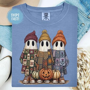 May include: A blue t-shirt featuring a graphic of three ghosts wearing beanies. The left ghost wears a maroon beanie and a mushroom-patterned sheet. The centre ghost wears a yellow beanie and a Celtic-patterned sheet. The right ghost wears an orange beanie and a thistle-patterned sheet. A jack-o'-lantern sits at their feet.