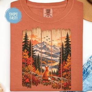 Peut inclure: Un t-shirt couleur terre cuite avec une impression graphique représentant un renard assis sur un rocher surplombant un lac, des montagnes et une forêt aux couleurs automnales. Le motif est encadré par des lattes de bois et porte une étiquette Comfort Colors.