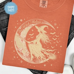 Può includere: Una t-shirt color terracotta con una grafica color crema raffigurante una strega su una scopa davanti a una luna crescente. Il testo "The Witch Melissa" è arcuato sopra la luna, circondato da piccole stelle e pipistrelli in volo.