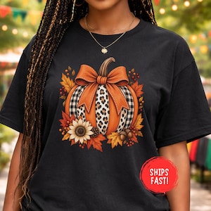 Könnte beinhalten: Ein schwarzes Kurzarm-T-Shirt mit einer zentralen Grafik eines Kürbisses, der mit einer großen orangefarbenen Schleife verziert ist. Der Kürbis hat abwechselnde Felder in Orange, schwarz-weißem Karomuster und Leopardenmuster, umgeben von Herbstblättern.