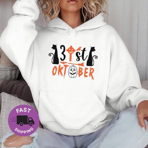 May include: White Halloween hoodie with the text "13st OKTOBER" in black and orange. The design includes black cat silhouettes, a pumpkin, a broom, and a cross. A purple circle with the words "FAST SHIPPING" is in the lower left corner.
