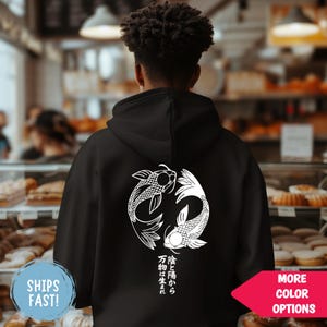 Könnte beinhalten: Schwarzer Kapuzenpullover mit weißem Yin-und-Yang-Koi-Fisch-Design auf dem Rücken, einschließlich japanischem Text. Der Hoodie hat eine Kapuze und lange Ärmel. Das Bild enthält auch eine Schaltfläche "MORE COLOR OPTIONS".